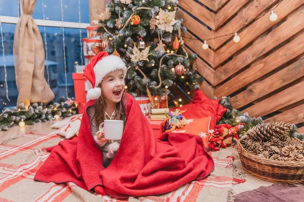 Bellissimo bambino in cappello di Babbo Natale, avvolto in una coperta rossa vicino all'albero di Natale, con una tazza di cacao, dall'aspetto felice e soddisfatto . — Foto Stock