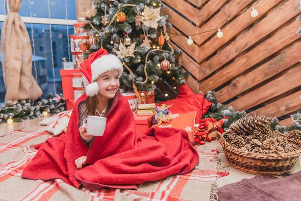 Bellissimo bambino in cappello di Babbo Natale, avvolto in una coperta rossa vicino all'albero di Natale, con una tazza di cacao, dall'aspetto felice e soddisfatto . — Foto Stock