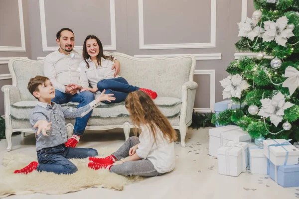 Niño y niña conjurando copos de nieve con las manos, mientras los padres los miran, sentados en el sofá . — Foto de Stock