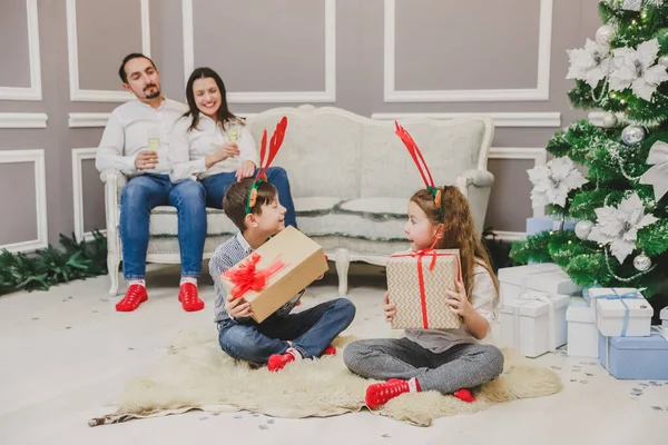 Foto de cuerpo completo de niños lindos sentados en posición lotos con un regalo de Navidad en las manos, averiguando lo que hay dentro, mientras los padres están sentados en el sofá, bebiendo champange . —  Fotos de Stock