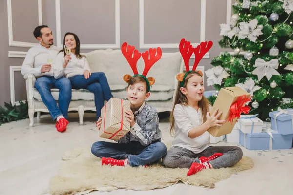 Foto de cuerpo completo de niños lindos sentados en posición lotos con un regalo de Navidad en las manos, averiguando lo que hay dentro, mientras los padres están sentados en el sofá, bebiendo champange . —  Fotos de Stock