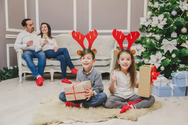 Fratelli felici con divertenti corna di renna rosse sulla testa e enormi scatole regalo sono seduti fianco a fianco pronti ad aprire i regali. Sfondo sfocato dove madre e padre si godono il momento . — Foto Stock