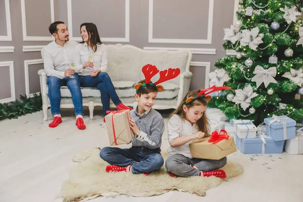Szczęśliwe rodzeństwo ze śmiesznymi czerwonymi rogami reniferów na głowie i ogromnymi pudełkami prezentów siedzą obok siebie gotowi do otwarcia prezentów. Zamazane tło, w którym rodzice cieszą się chwilą. — Zdjęcie stockowe