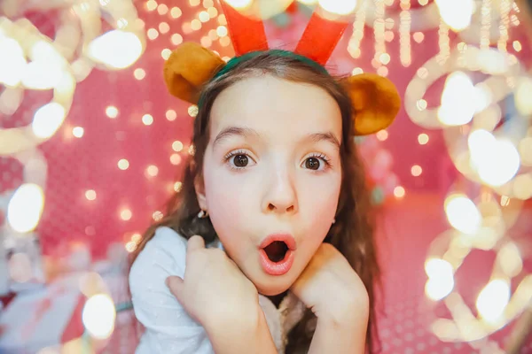 Bokeh-Lichter aus einer Weihnachtsgirlande auf der Vorderseite und lustiges kleines Mädchen mit Rentiergeweih, das in die Kamera schaut, abends zu Hause lächelt. — Stockfoto