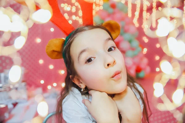Bokeh-Lichter aus einer Weihnachtsgirlande auf der Vorderseite und lustiges kleines Mädchen mit Rentiergeweih, das in die Kamera schaut, abends zu Hause lächelt. — Stockfoto