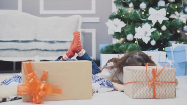 Sorglose, lächelnde kleine Geschwister genießen die Weihnachtszeit, liegen auf dem Teppich unter dem Tannenbaum und zeigen ihre Köpfe hinter zwei Geschenkboxen. — Stockvideo