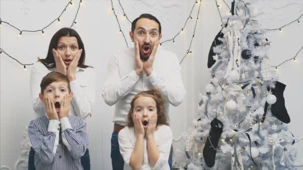 4k video van jonge ouders en hun kinderen op nieuwjaarsachtergrond, met monden wijd open en handen op de wangen, kijkend naar de camera met horror in de ogen. — Stockvideo