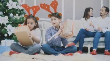 Ellerinde Noel hediyesiyle loto pozisyonunda oturan ve içinde ne olduğunu öğrenen sevimli çocukların tüm vücudunun videosu. Aileler kanepede oturmuş şampanya içerken..