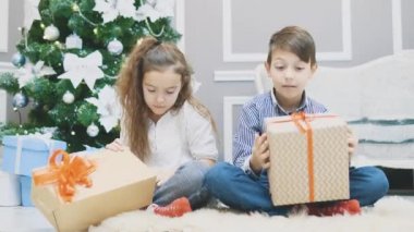 Sevimli erkek ve kız kardeşler loto pozisyonunda ellerinde Noel hediyeleriyle oturup, onu çalkalayıp, çalkalayıp, içindekileri dinliyorlar..