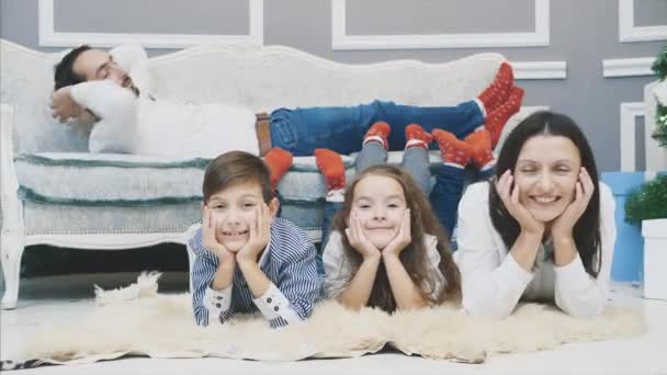 Two kids and mother putting legs on the sofa, elbows on the floor, looking at the camera and father laying on the sofa upside-down. All are plaing a game in holiday atmosphere. — Stock Video