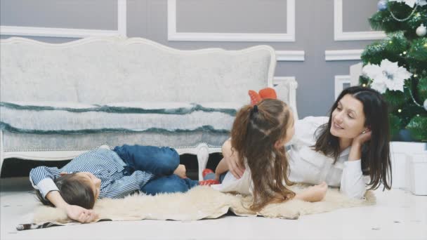 Niña y niño con su madre están sentados en la alfombra de piel cerca de abeto, disfrutando de tiempo juntos . — Vídeos de Stock