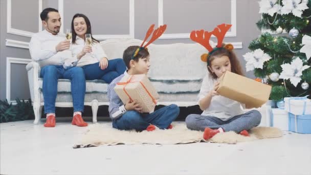 Helkroppsvideo av söta barn som sitter i lotos position med en julklapp i händerna, ta reda på vad som finns inuti, medan föräldrarna sitter på soffan, dricka champange. — Stockvideo