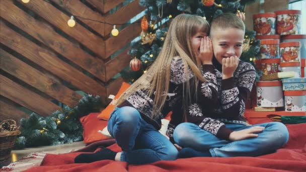 Süße Kinder flüstern dem einen oder anderen Geheimnisse ins Ohr, sitzen zu Hause auf einer roten Decke und feiern Weihnachten. — Stockvideo