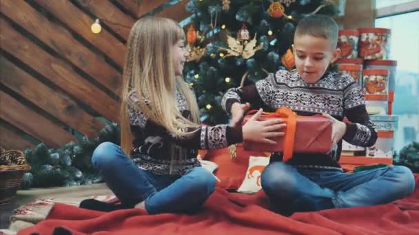 Lovely 4k vídeo of sister giving christmas gift box to her brother while sitting near tree, in christmas designed atmosphere . — Vídeo de Stock