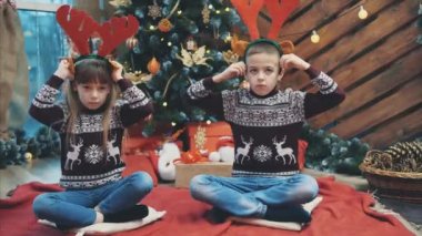 Ren geyiği boynuzu takan erkek ve kız kardeşlerin Noel akşamı köknar ağacında eğlendikleri 4k sevimli bir video..