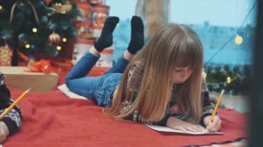 Karnının üzerine uzanmış, Noel Baba 'ya mektup yazan şirin, küçük bir çocuk. Arka planda ışıklar ve hediyeler olan Noel ağacı.