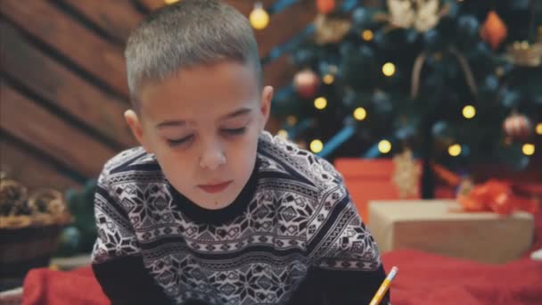 Милый мальчик в праздничном платье лежит на полу и пишет письмо Санте. Рождественская елка с огнями и подарками на заднем плане . — стоковое видео