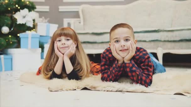 Hermano sonriente y hermana pequeña en previsión de año nuevo están acostados en la alfombra de lana, vestidos con ropa de cuadros rojos . — Vídeos de Stock
