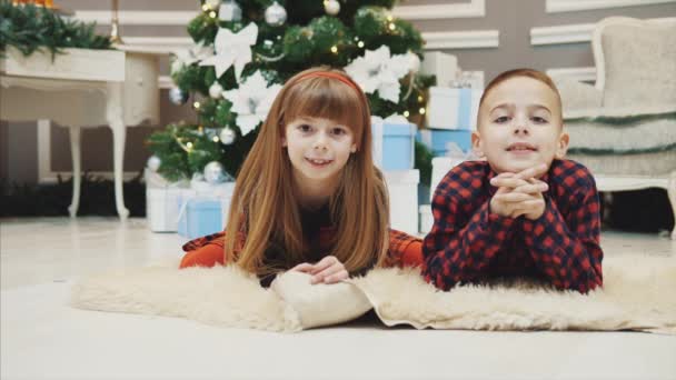 Usmívající se bratr a sestřička v očekávání nového roku leží na vlněném koberci v červených kostkovaných šatech. — Stock video