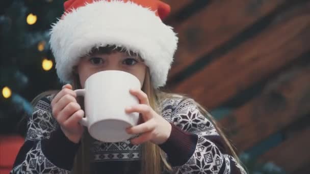 Close-up 4k video van schattig lachend meisje zitten gezellig thuis, met een kopje warme chocolademelk bij kerst boom op houten achtergrond. — Stockvideo