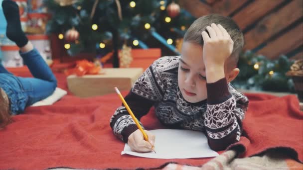 Aranyos kisfiú, ünnepi ruhában fekszik a földön, levelet ír a Mikulásnak. Karácsonyfa fényekkel és ajándékokkal a háttérben. — Stock videók