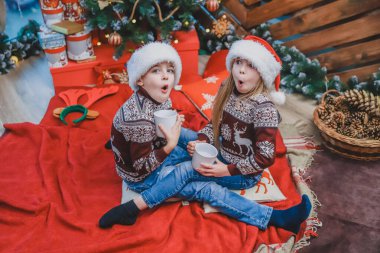 Up-view shot of amazed children in santa hats and woolen sweaters, sitting near christmas tree, drinking cocoa, looking up, their mouths widely opened. clipart