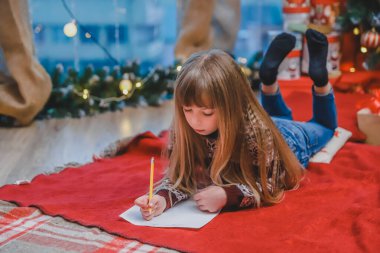 Yerde yatan, Noel Baba 'ya mektup yazan, şenlikli elbiseli şirin bir çocuk. Arka planda ışıklar ve hediyeler olan Noel ağacı.