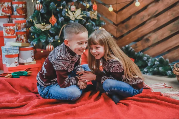 Dolce scatto di un ragazzo che racconta un segreto nell'orecchio a sua sorella seduta su una coperta rossa a casa, mentre festeggia il Natale . — Foto Stock