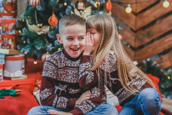 Dulce foto de una chica contando un secreto al oído a su hermano sentado en una manta roja en casa, celebrando la Navidad . — Foto de Stock