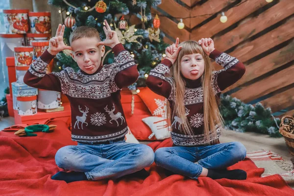 I bambini sorridenti in maglioni di lana mostrano grandi orecchie con le mani e mettono lingue su sfondo di legno con albero di Natale . — Foto Stock