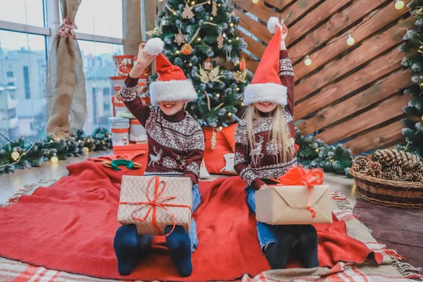 Foto de hermanos en jerséis navideños con estampado de reno y con cajas en las piernas, burlándose, poniéndose sombreros de santa en los ojos y subiéndolos . — Foto de Stock