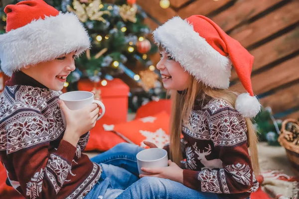 Крупним планом фото розумних дітей, які носять капелюхи Санти напередодні Різдва, п'ють гаряче какао і цікавляться, що вони отримають в подарунок . — стокове фото