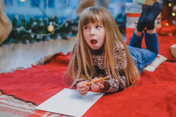 Lindo niño con vestido festivo tirado en el suelo, escribiendo una carta a Santa. Árbol de Navidad con luces y regalos al fondo . — Foto de Stock