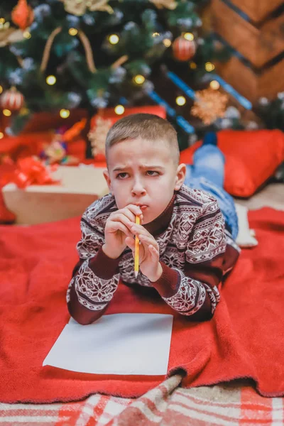 Criança bonito em vestido festivo deitado no chão, escrevendo carta para o Papai Noel. Árvore de Natal com luzes e presentes no fundo . — Fotografia de Stock