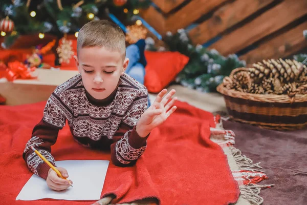 Criança bonito em vestido festivo deitado no chão, escrevendo carta para o Papai Noel. Árvore de Natal com luzes e presentes no fundo . — Fotografia de Stock