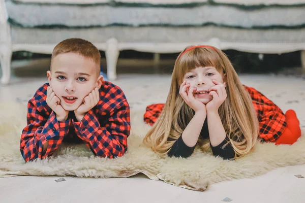 Irmão sorridente e irmã mais nova estão deitados no tapete de lã, vestidos com roupas xadrez vermelho . — Fotografia de Stock