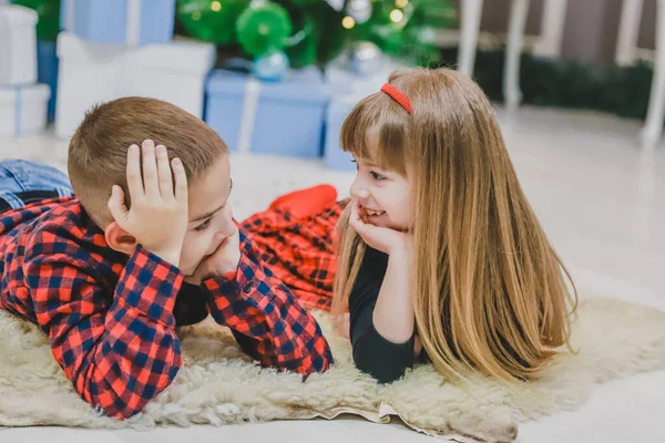 Hermano sonriente y hermana pequeña están acostados en la alfombra de lana, vestidos con ropa de cuadros rojos . — Foto de Stock