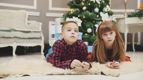 Foto cálida de los hermanos bonitos viendo una película de año nuevo mientras se ponen en la alfombra bajo el abeto de Navidad . — Vídeos de Stock