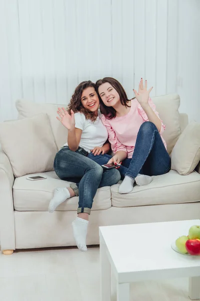Dvě úžasné dívky sedící na gauči, objímající se, mávající na sebe a usmívající se do kamery. — Stock fotografie