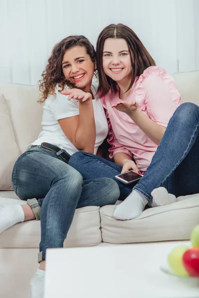 Zwei erstaunliche Mädchen, die auf dem Sofa sitzen, einander umarmen, Luftküsse geben und in die Kamera lächeln. — Stockfoto