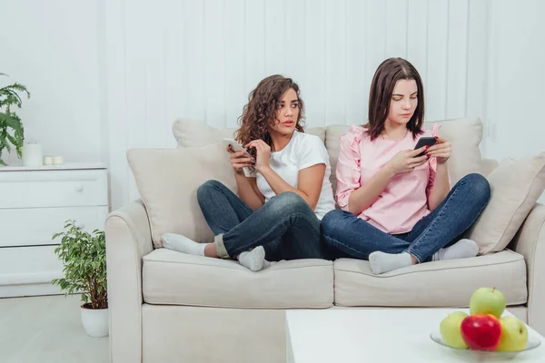 Zwei attraktive Freundinnen, die nebeneinander auf dem Sofa in Lotos-Position sitzen, mit Smartphones in der Hand. — Stockfoto