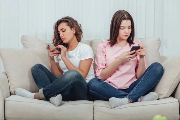 Zwei attraktive Freundinnen, die nebeneinander auf dem Sofa in Lotos-Position sitzen, mit Smartphones in der Hand. — Stockfoto
