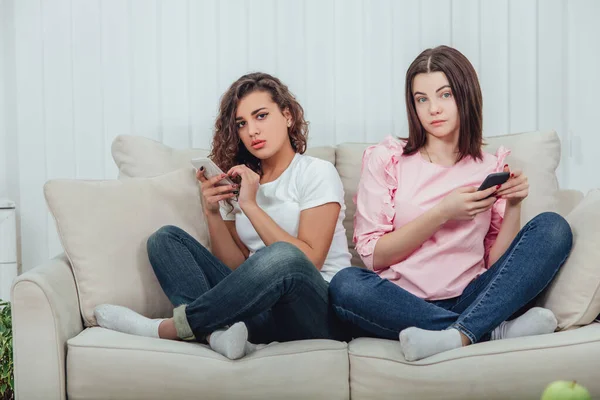Zwei attraktive Freundinnen, die nebeneinander auf dem Sofa in Lotos-Position sitzen, Smartphones in der Hand und in die Kamera schauen. — Stockfoto