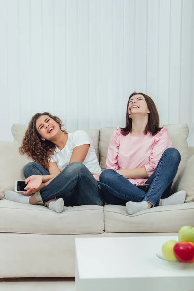 Due ragazze attraenti sedute sul divano in posizione foto, con gli smartphone in mano, sorridendo disperatamente . — Foto Stock