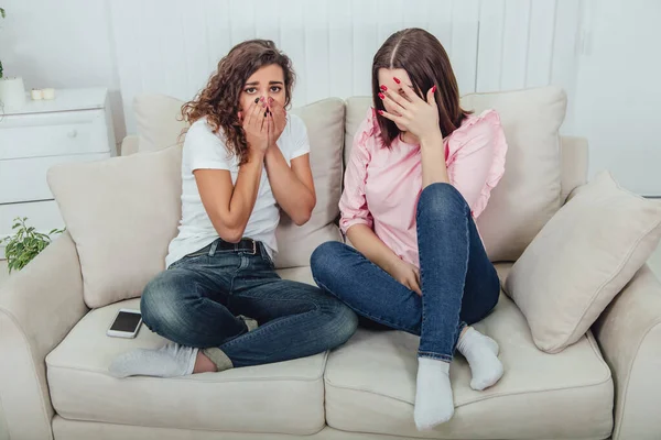 Two attractive girlfriends sitting on the sofa discussing something that evokes strong emotions. Surprised even scared face expressions. — Stock Photo, Image
