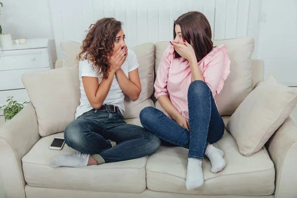 Dvě atraktivní přítelkyně sedící na gauči a diskutující o něčem, co vyvolává silné emoce. Překvapen i vyděšené výrazy obličeje. — Stock fotografie