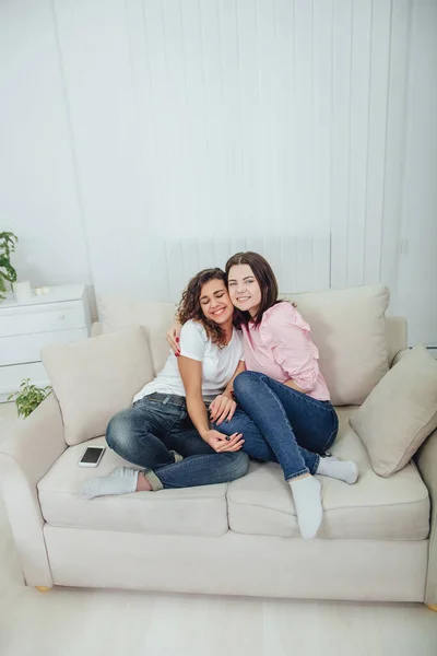 Дві дивовижні дівчата сидять на дивані, обіймаючи один одного, усміхаючись мило, дивлячись на камеру . — стокове фото