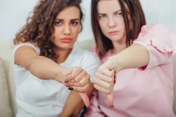 Blurred cute girls sitting on the sofa side by side, giving thumbs down, frowning. Focus on the thumbs on the forefront. — Stock Photo, Image