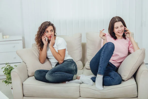 Due ragazze fantastiche sedute sui diversi lati del divano, che parlano al telefono. Una ragazza sta parlando forte e apertamente, un'altra sta tenendo la mano vicino alla bocca, parlando piano . — Foto Stock