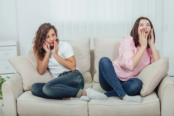 Dos chicas increíbles sentadas en los diferentes lados del sofá, hablando en los teléfonos. Una chica habla fuerte y abiertamente, otra mantiene la mano cerca de la boca, hablando en voz baja . — Foto de Stock